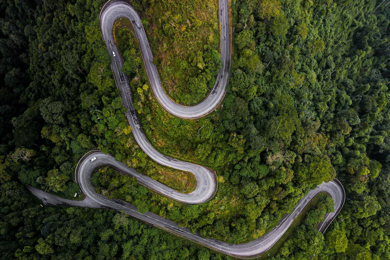 gallery-4 Drone shot of a serpentine road cutting through a lush green mountainous forest landscape.