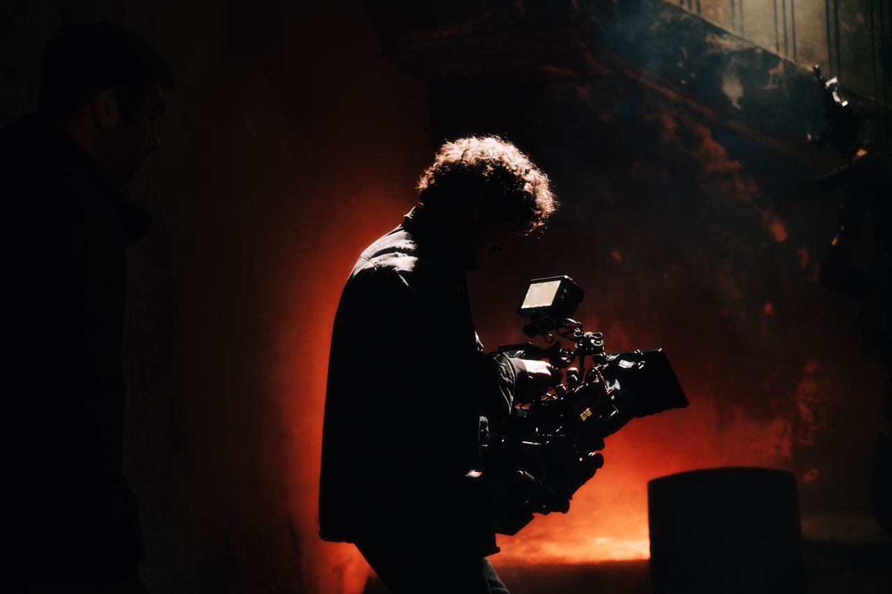 our-services-1 A professional cameraman captures footage in a creatively lit, moody setting, backlit by warm light.